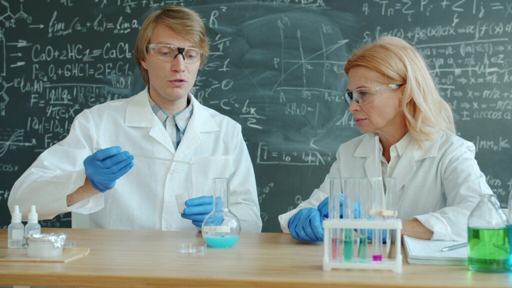 Two scientists in lab coats and gloves preparing chemicals at a laboratory bench